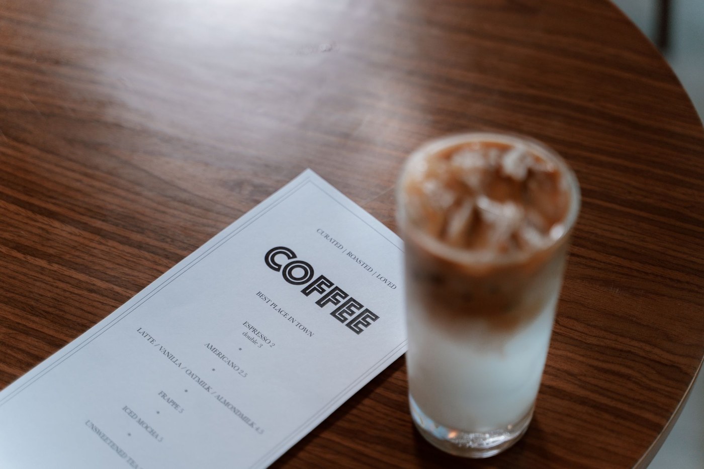 An iced coffee beside a printed English coffee menu on a wooden table